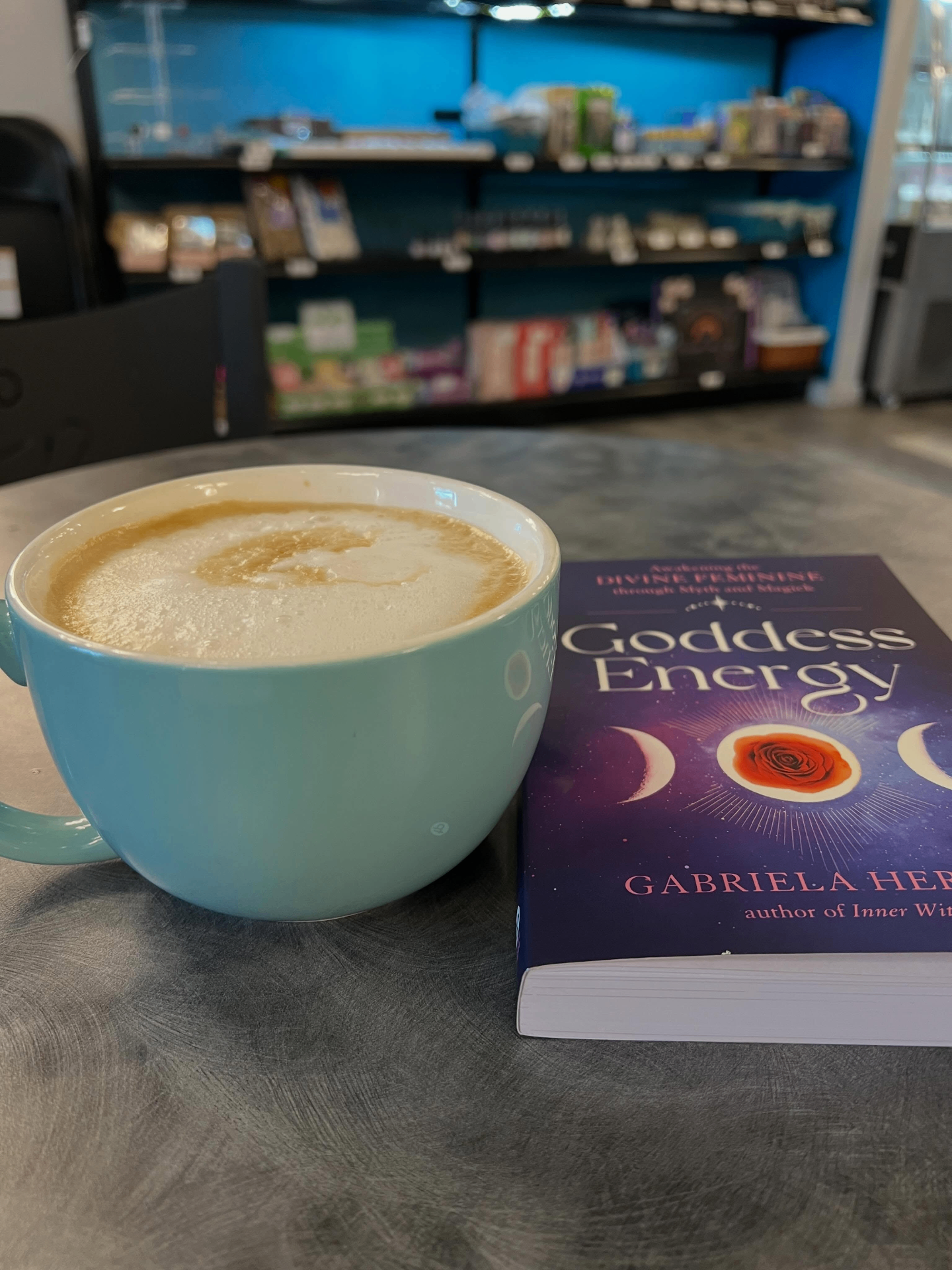 Light blue latte mug beside the Goddess Energy book on a cafe table at Something Wicked Coffee